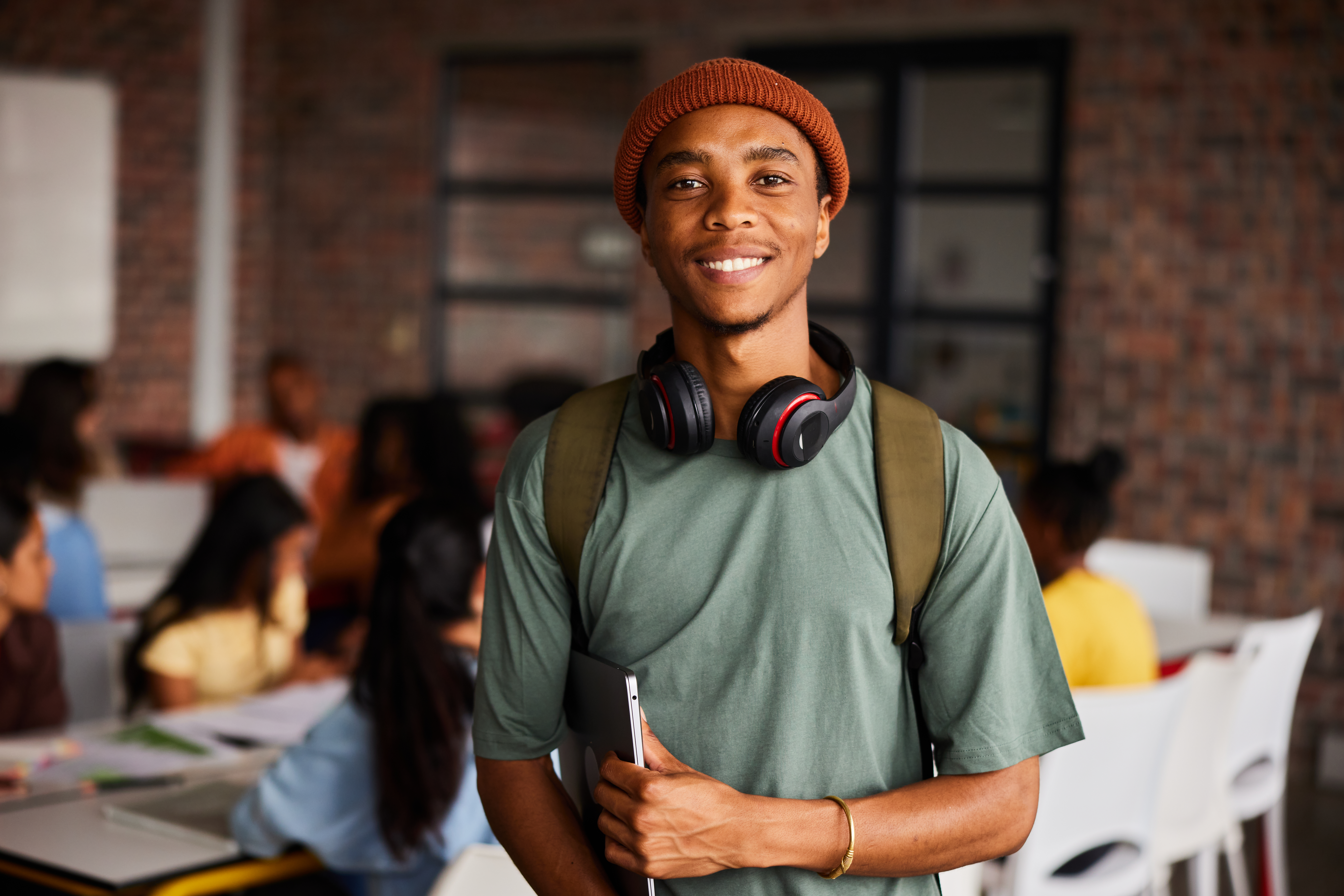 Portrait of a young male college student wearing headphones and a beanie smiling while standing in a classroom with students behind him